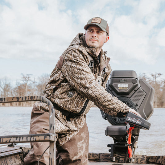 A man in Guardian Elite HND Front Zip Waders, standing confidently on a boat. Waterproof, breathable 4-layer upper body and 5-layer legs for ultimate protection. Ideal for hunting in various terrains.