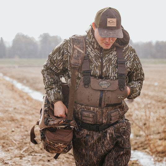 A man in the field with hat and bag, showcasing the Buckshot Eqwader 1600 Neoprene Wader 3.0 by Drake Waterfowl, designed for top-tier waterfowl hunting with advanced features for comfort and durability.