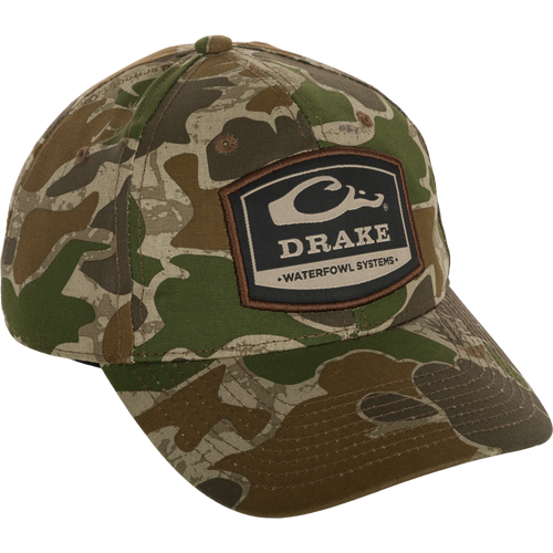 A 6-Panel Badge Cap with a camouflage pattern and a patch/logo on it. Made of cotton blend panels, this cap features a secure fit with a rear snap closure. Perfect for outdoor enthusiasts.