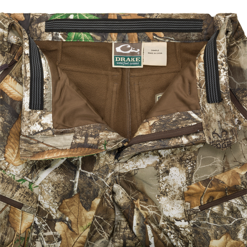 MST Microfleece Softshell Pant - Realtree: Close-up of camouflage pants with zipper, label, and chain. Superior protection and comfort with wind-resistant fabric, 4-way stretch, and moisture-repellent lining. Scent control technology, comfortable fit, and easy movement with articulated knees and side zips. Zippered pockets for storage.