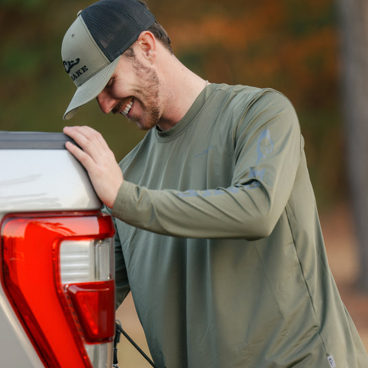 A man in a hat smiles, showcasing the L/S Performance Crew Solid by Drake Waterfowl. Features include UPF 50, moisture-wicking, and quick-drying fabric for outdoor activities.