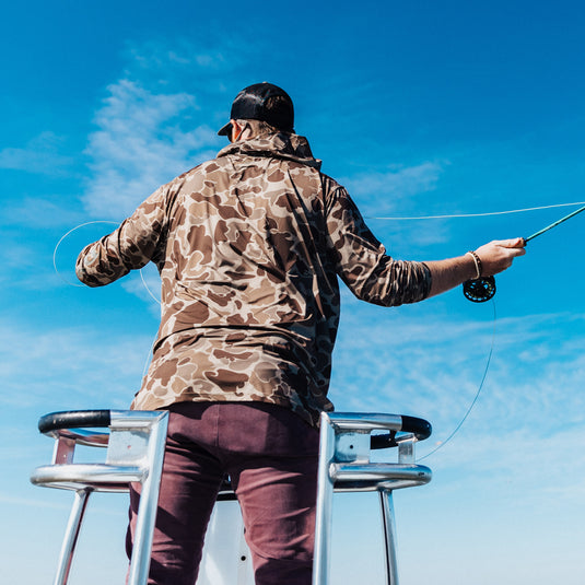 A man on a boat holding a fishing pole, wearing the Hunter Creek Old School Bamboo Long Sleeve Hoodie by Drake Waterfowl. Lightweight, odor-resistant, with UPF 20 sun protection.