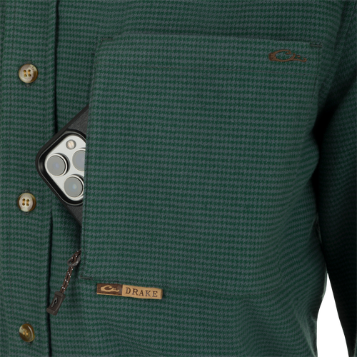 A person wearing a green jacket with a cell phone in it, close-up of button, camera lens, label, and zipper. Autumn Brushed Twill Houndstooth Shirt.