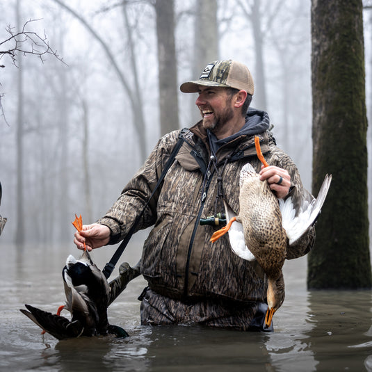 A man in a versatile Reflex™ 3-IN-1 Plus 2 Jacket, holding ducks in the water. Waterproof, windproof, and breathable with multiple pockets and adjustable features for hunting comfort.