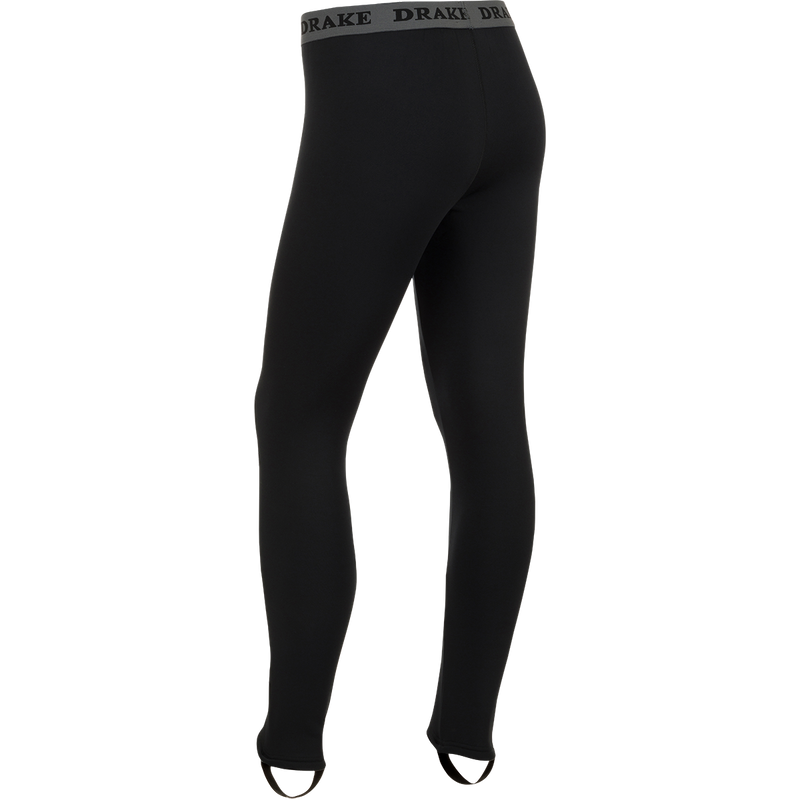 LST Heavyweight Baselayer Pant: A person's legs in black tights wearing a black pants with a grey band. Ideal for active and cold weather activities. Exceptional stretch, extreme warmth, and moisture-wicking properties. Stirrup strap for layering under waders and front fly for convenience. Stay warm and comfortable in any condition.