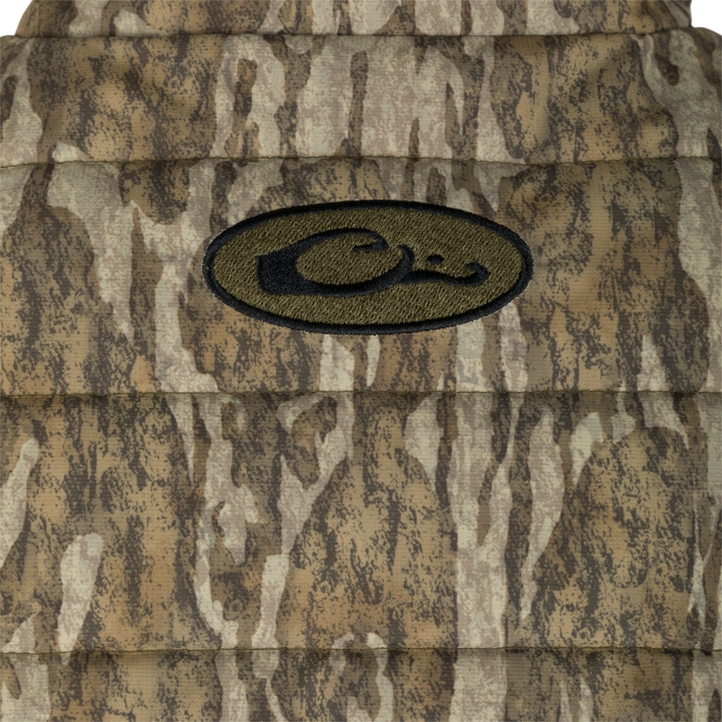 A close-up of the LST Double Down Layering Full Zip vest, featuring a logo on a camouflage pattern. Stay warm in style with this insulated vest made of 100% Polyester Synthetic Down. Updated fit and reverse-coil zippers provide comfort and functionality. Zippered pockets and adjustable waist for convenience. From Drake Waterfowl, your go-to for high-quality hunting gear and clothing.