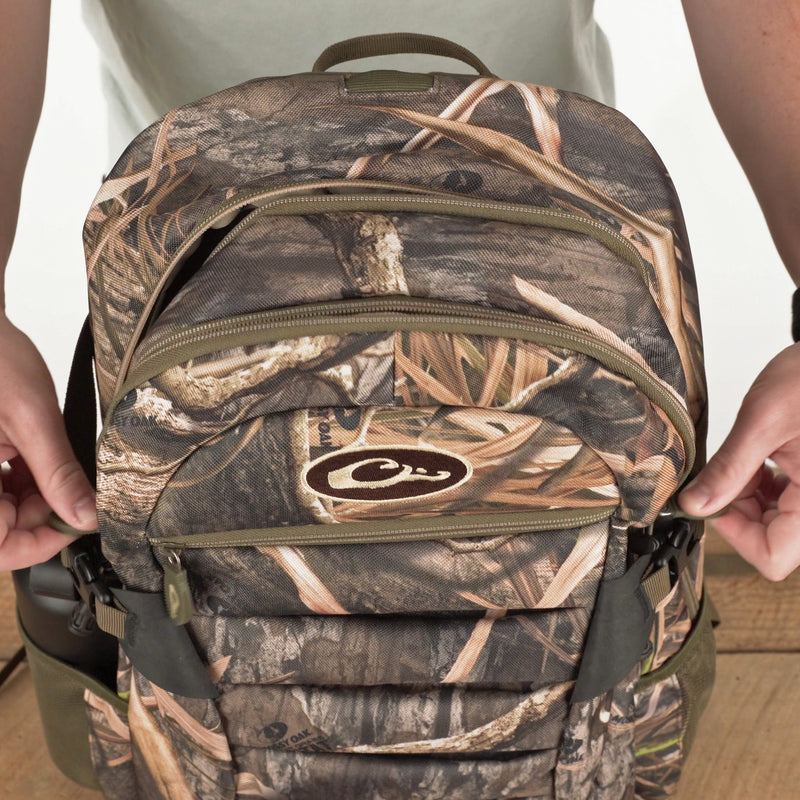 A rugged Camo Daypack by Drake Waterfowl, ideal for hunting and outdoor adventures. Features include water-resistant material, padded backing, ample storage, hydration pouch, and adjustable straps. Dimensions: 18.5 x 11.5 x 8.5.