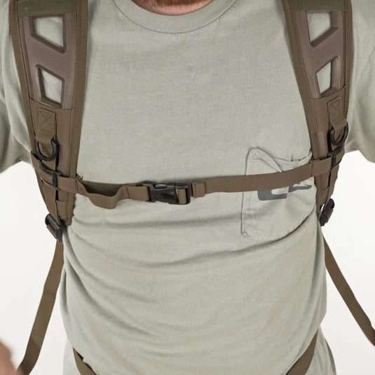 A man wearing a grey shirt and grey pants, showcasing the Camo Daypack by Drake Waterfowl. Features include a large storage area, hydration pouch pocket, EVA shoulder straps, and more.