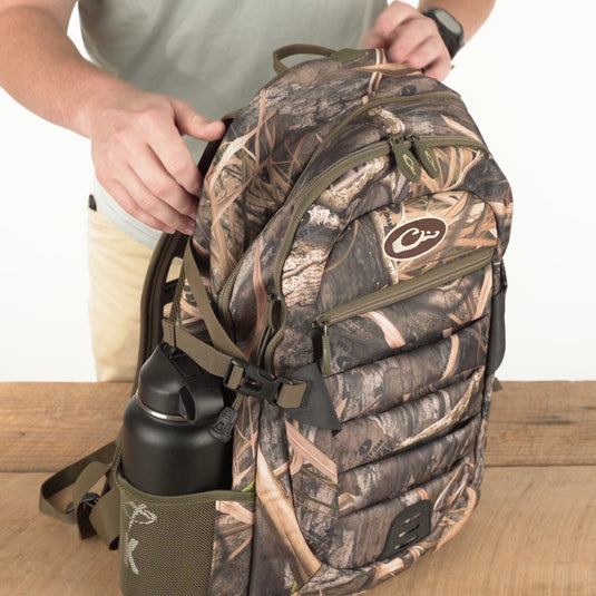 A rugged Camo Daypack by Drake Waterfowl, ideal for versatile use from classroom to field. Features include ample storage, hydration pockets, padded backing, and durable HD2™ material. Dimensions: 18.5 x 11.5 x 8.5.