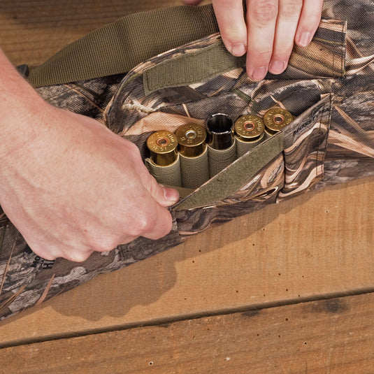 A side-opening gun case with a hand holding a bag of cartridges and bullets inside. Rugged HD2™ material exterior with a nylon interior liner. Features adjustable strap, top carry handle, and quick-top hook & loop opening. Accommodates shotguns up to 52" overall length.