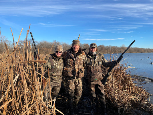 A group of men in camouflage standing in a wetland area, wearing camouflage clothing and holding guns.