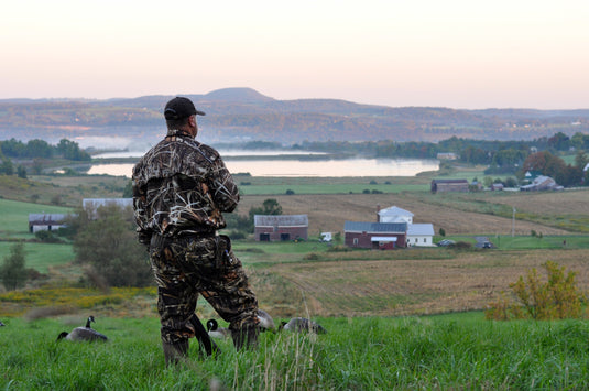 A man in camouflage outfit standing in a field, looking at a lake.