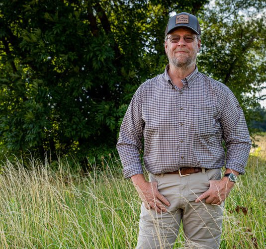 A man standing in a field wearing a plaid shirt, hat, and glasses, holding his hands on his hips.