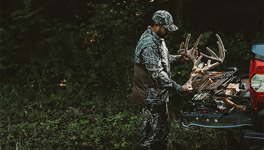 A man in camouflage holding the deer's head, with a deer on the back of a pickup
