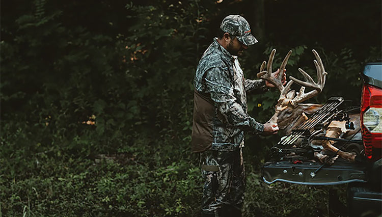 A man in camouflage holding the deer's head, with a deer on the back of a pickup