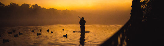Silhouetted hunter with a shotgun in calm water at dawn, surrounded by duck decoys, reflecting Drake Waterfowl's commitment to quality hunting gear.