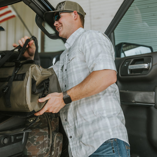 A man in sunglasses and a hat holding a bag near a vehicle, showcasing the Frat Faded Plaid Button-Down Short Sleeve Shirt from Drake Waterfowl. Features UPF 30, moisture-wicking, and hidden collar.