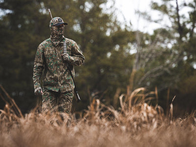 Man in camouflage holding a gun, wearing the Throwback 80 Button Down Field Shirt with reinforced forearms, ideal for outdoor hunting adventures.