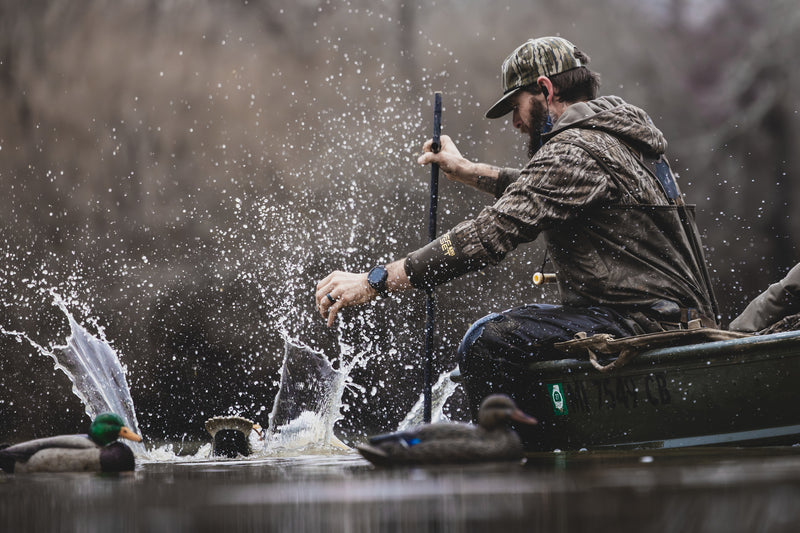 Man in camo waders leans over a boat with duck decoys nearby, wearing BMF EST Offset Quarter Zip Hoodie, ideal for waterfowl hunting with advanced features.