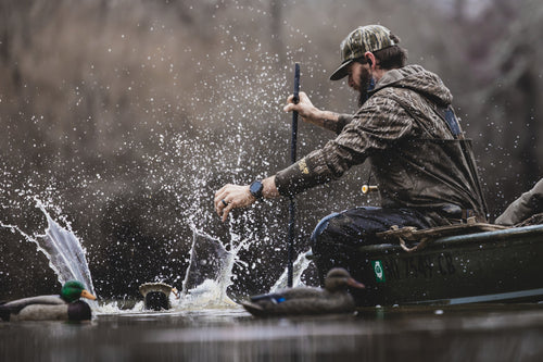 Man in camo waders leans over a boat with duck decoys nearby, wearing BMF EST Offset Quarter Zip Hoodie, ideal for waterfowl hunting with advanced features.