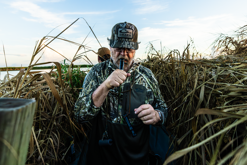 Hunter in camouflage using a duck call, showcasing the EST Performance Half-Zip Hoodie with ergonomic thumbholes and water-resistant pocket in a wetland hunting setup.