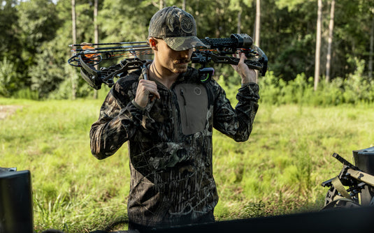 A person in camo stands outdoors with a compound bow, wearing the Nontypical Non-Typical Lightweight SWT Half Zip Pullover with Scent Control amid green grass and trees.