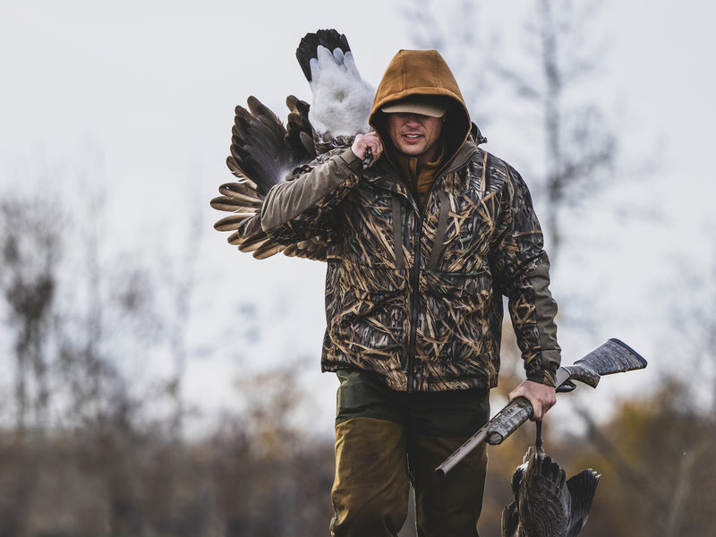 Hunter in camouflage with the LST BMF 3-in-1 Pit Jacket, carrying geese and a shotgun, showcasing durable, versatile hunting gear from Drake Waterfowl.