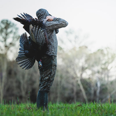 A man outdoors wearing a Turkey Bib with Spider Web Technology, featuring stretch material, cargo pockets, and water-resistant knee reinforcement, while carrying a turkey on his back.