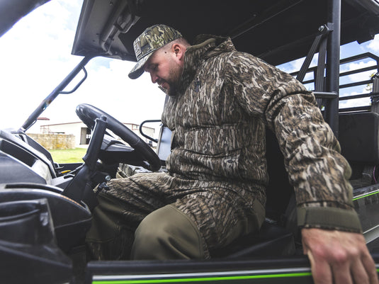 A man in a camouflage Spoonie Hollywood Pant, jacket, and cap sits in the driver’s seat of an all-terrain vehicle, wearing wind-resistant hunting apparel.