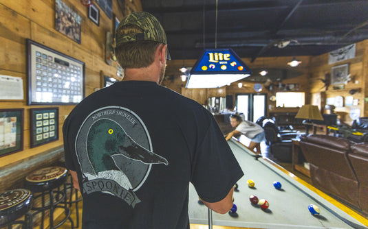 A person standing by a pool table wearing a black Spoonie Head Logo T-Shirt with a graphic of a Northern Shoveler duck head on the back.