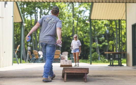 A man in a grey Spoonie Hollywood Logo T-Shirt and jeans prepares to throw a bean bag during a cornhole game, while a woman waits in the background.