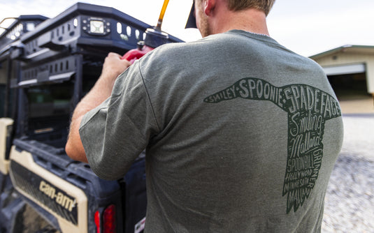 A man wears an olive green Spoonie Spade Face Logo T-Shirt with a back graphic shaped like a duck head containing the words Spoonie, Spade Face, Smiling Mallard, Spoonbill, Hollywood, Minnesota, and Mallard.
