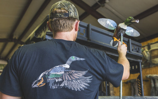 A person wearing a black Spoonie In-Flight Logo T-Shirt with a detailed illustration of a flying shoveler duck on the back.