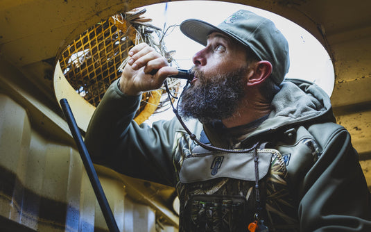 A bearded man wearing a camo Spoonie Hollywood Jacket and cap blows into a duck call while looking upward from a blind.