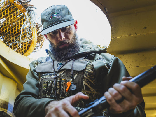 A bearded man in a camo hunting jacket and Spoonie Brand In Flight Cap looks down as he handles a shotgun.