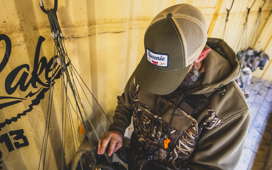 A man wearing a green and tan mesh-back Spoonie Banquet Cap with a rectangular patch on the front, looking down while handling thin cords.