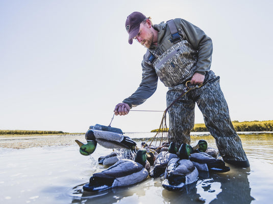 A hunter in camouflage Spoonie Hollywood Breathable Wader waders places a mallard duck decoy into the water.