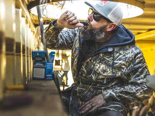 A bearded man wearing an insulated Spoonie Campfire Hoodie and a baseball cap uses a duck call inside a yellow blind.