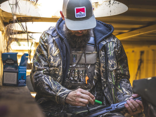 A bearded person wearing a grey and brown Spoonie Man Patch Cap and a camouflage jacket works on a shotgun inside a blind.