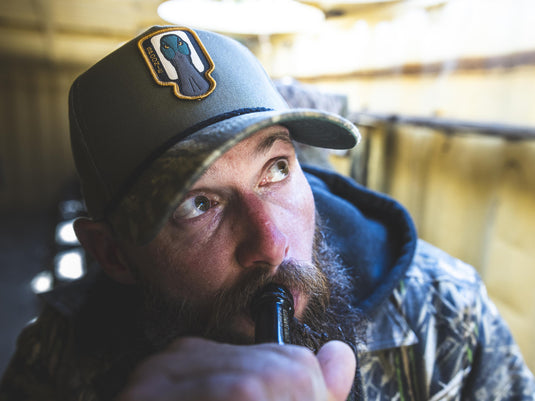 A bearded man wearing a Spoonie Logo Cap looks upward while using a duck call.