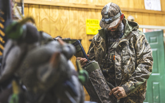 A man wearing a camo waterproof Spoonie Shoveler Insulated Jacket and headlamp packs a shotgun into a protective case.