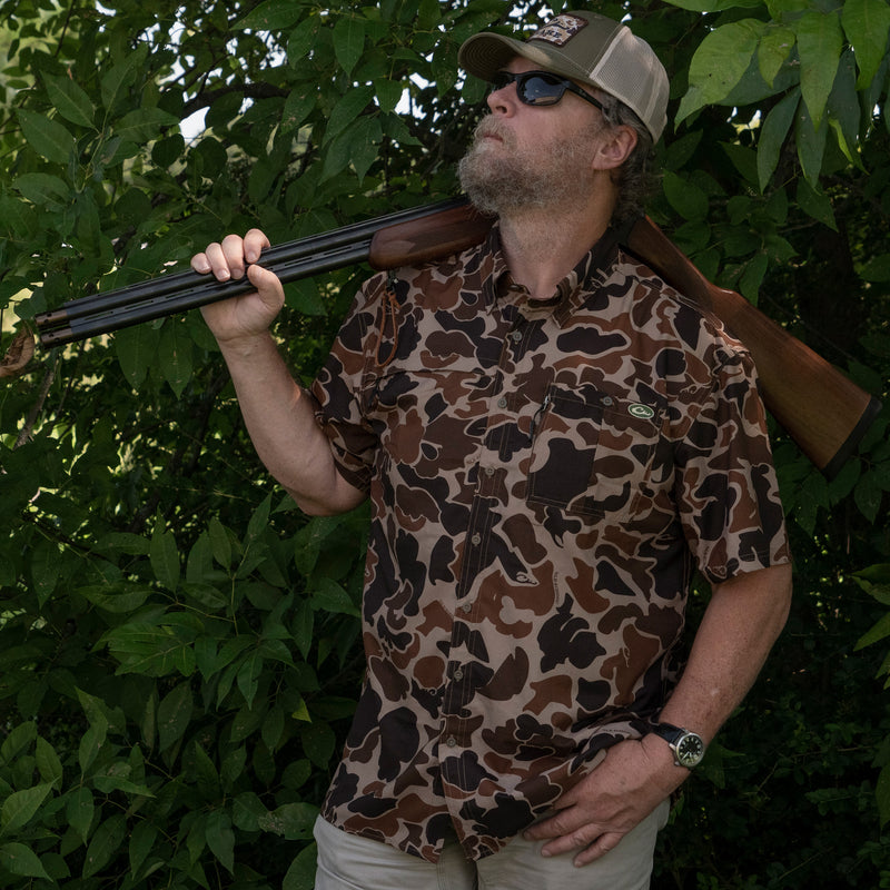 A man in camo holds a shotgun, wearing Drake 8 Shot Shirt. Lightweight, moisture-wicking fabric with hidden pockets and UPF30 sun protection. Ideal for hunting and outdoor activities.
