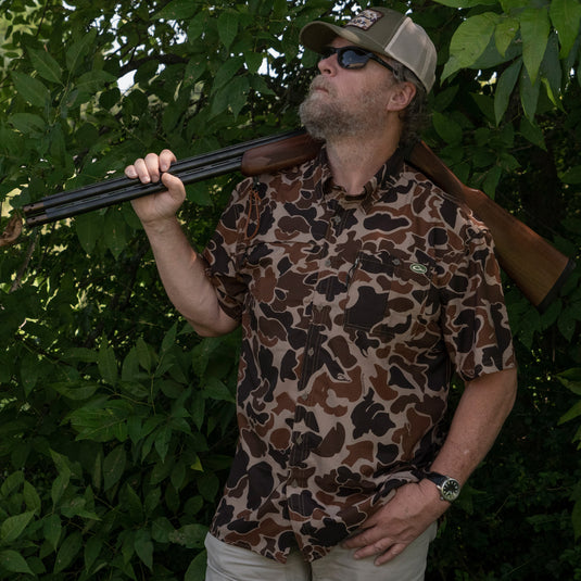 A man in camo holds a shotgun, wearing Drake 8 Shot Shirt. Lightweight, moisture-wicking fabric with hidden pockets and UPF30 sun protection. Ideal for hunting and outdoor activities.