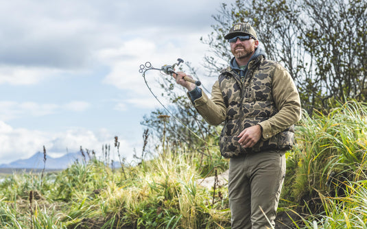 A man wearing a Waterfowl BMF Insulated Down Vest and camouflage cap holds a fishing rod while standing in tall grass.