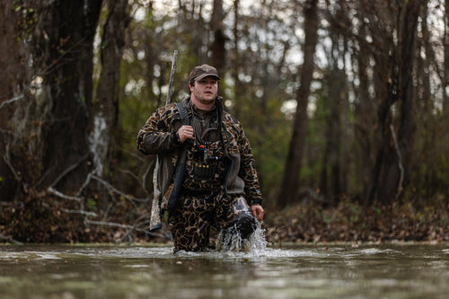 Person wearing MST CommandXT Eqwader Full Zip Jacket, walking in a flooded forest with a shotgun, showcasing the jacket's waterproof and durable features suitable for hunting.