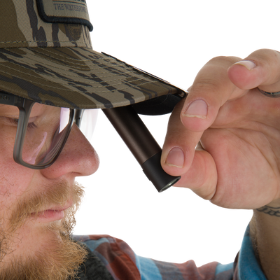 Man wearing a camouflage cap holds the EVO Cap Light under the brim, showcasing its compact design and hands-free functionality with a 360° pivoting clip.