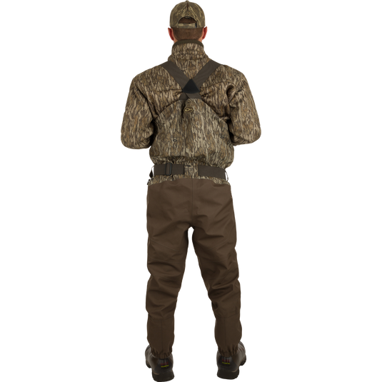 A man wearing camouflage clothing and a hat stands in the outdoors wearing Uninsulated Guardian Elite HND Front Zip Waders - Habitat.