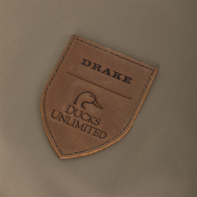 Shield-shaped leather patch featuring the embossed words Drake and Ducks Unlimited above a stylized duck silhouette, stitched onto olive-colored Kevlar fabric on the Drake X Ducks Unlimited BMF1600 Kevlar Wader.