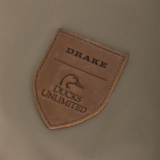 Shield-shaped leather patch featuring the embossed words Drake and Ducks Unlimited above a stylized duck silhouette, stitched onto olive-colored Kevlar fabric on the Drake X Ducks Unlimited BMF1600 Kevlar Wader.