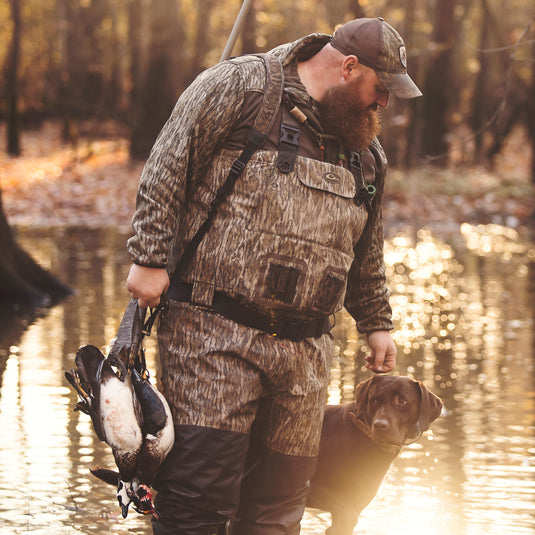 A man in camouflage holds a dog and a duck outdoors. Uninsulated Guardian Elite Vanguard Breathable Waders by Drake Waterfowl for extreme conditions, featuring waterproof fabric, reinforced seams, and Thinsulate Buckshot Mud Boot.